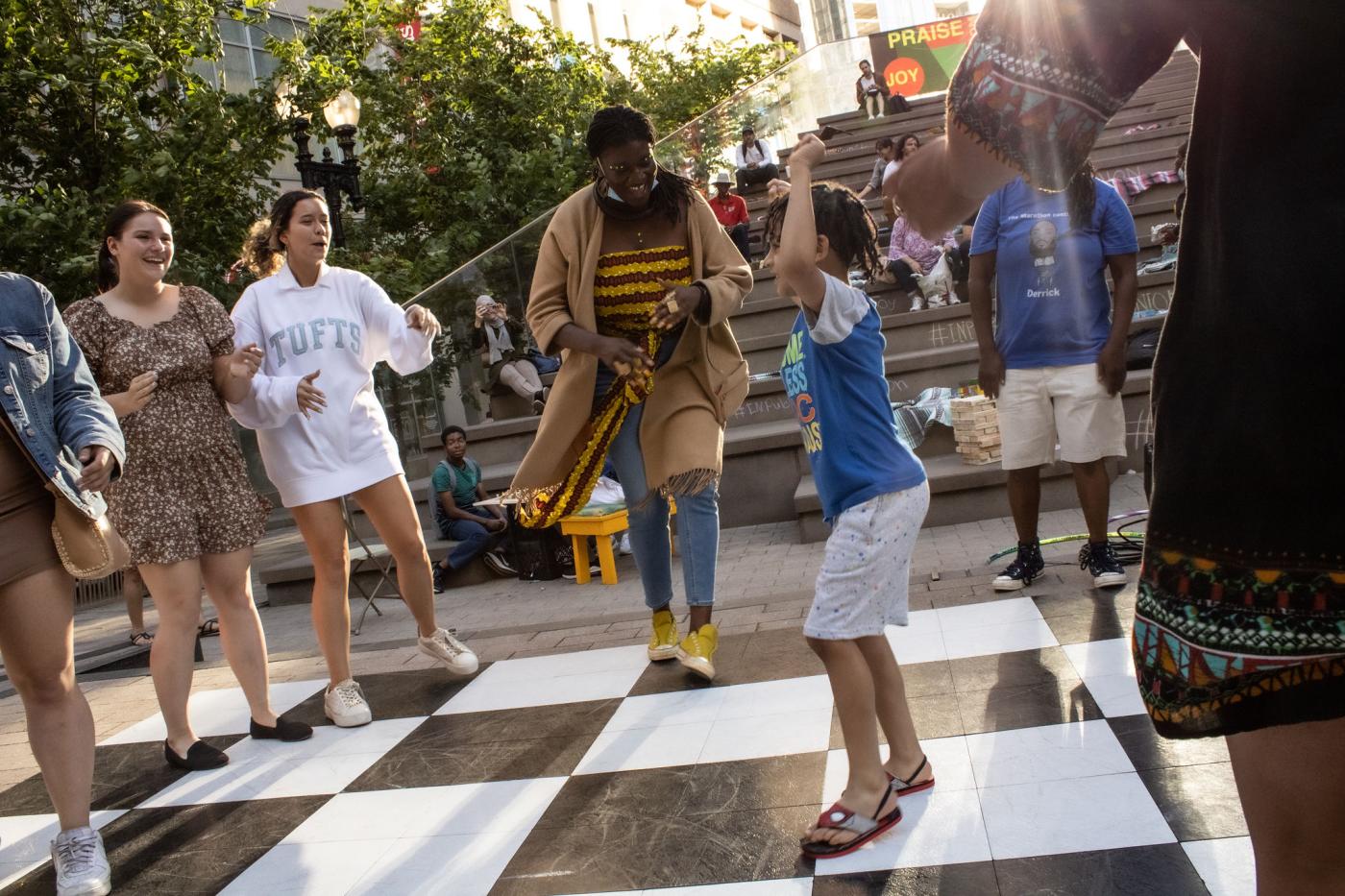 In a circle of folks, a young Black boy dances on top of a black and white checker board ground.