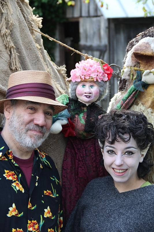 Outside, a white man, in a fedora, and a young white woman, with vampire ears and white powder on her face, pose with a doll or puppet.