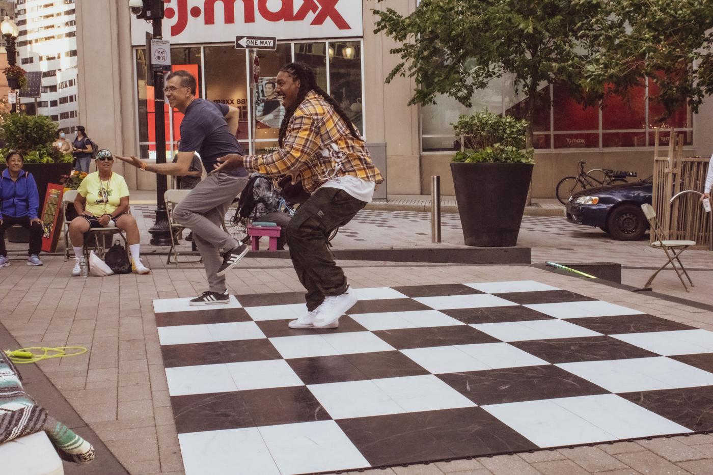 A light-skinned man and a dark-skinned person dance together on top of a checkerboard ground.