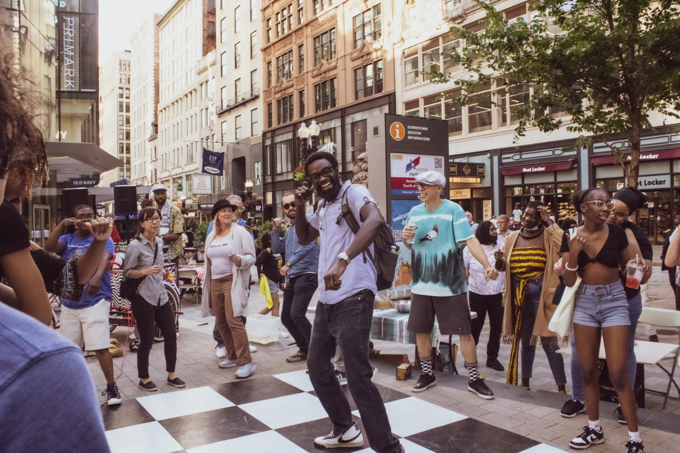 A crowd dances outside and, at the center of the crowd, a Black man with a short fro and a backpack smiles wide.