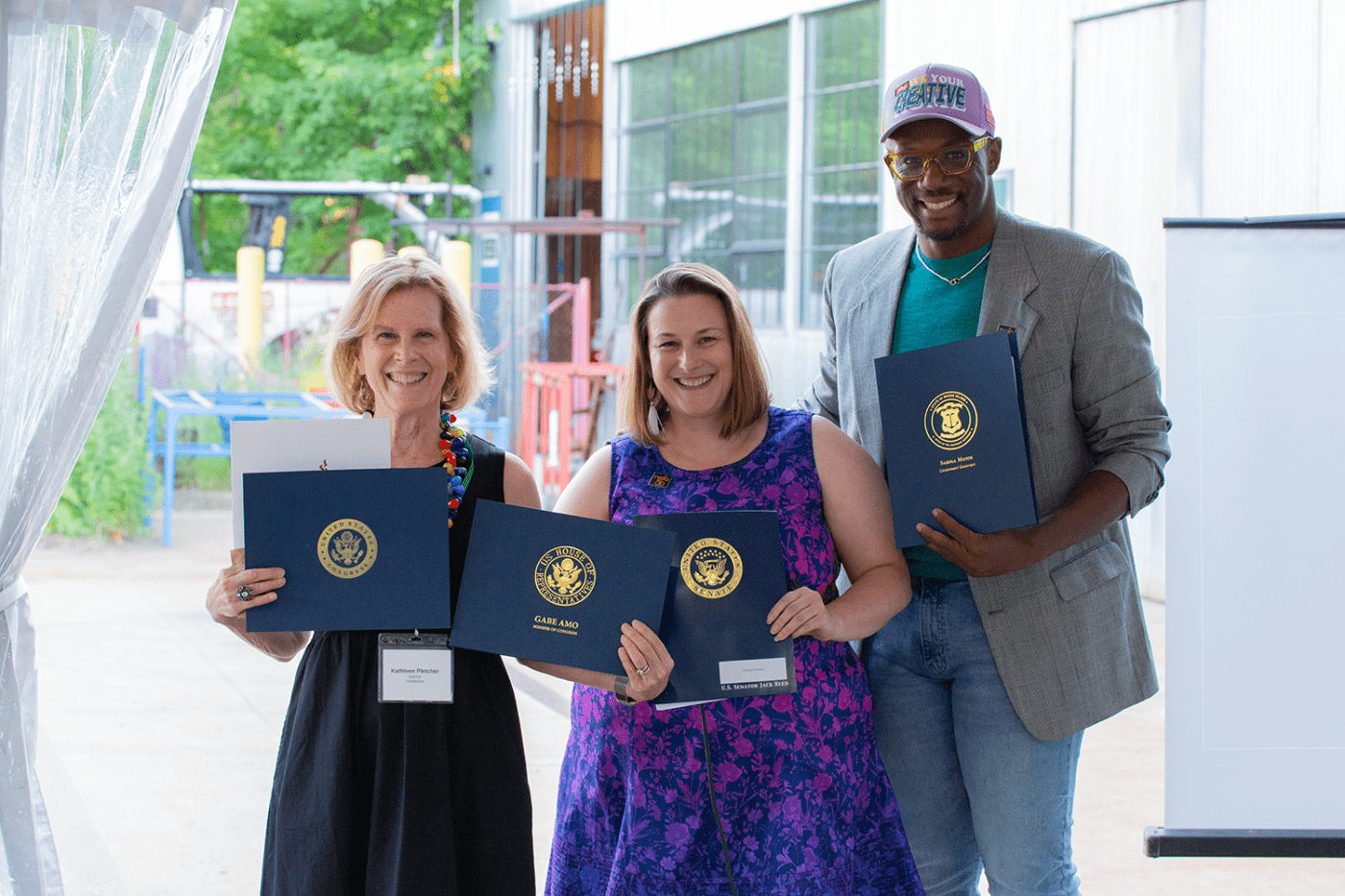 A blond white woman, in a black dress, poses with another white woman, in a purple and blue dress, and a Black man in a grey blazer and light-purple baseball cap.