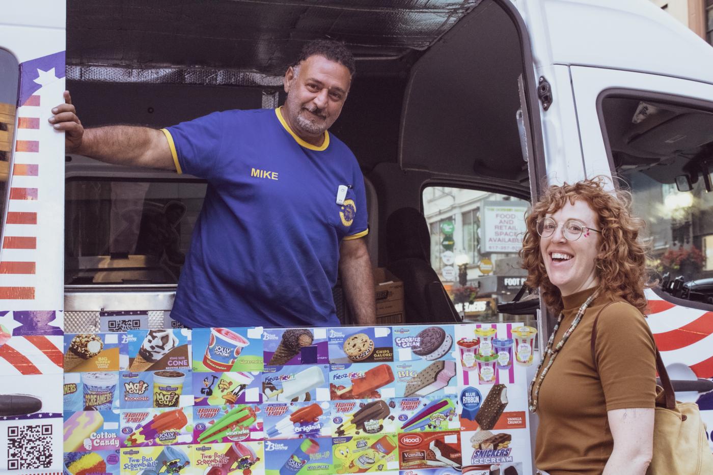 A man of color (his shirt says his name is Mike) in an ice cream truck poses with a white lady with red hair, who stands outside the truck.