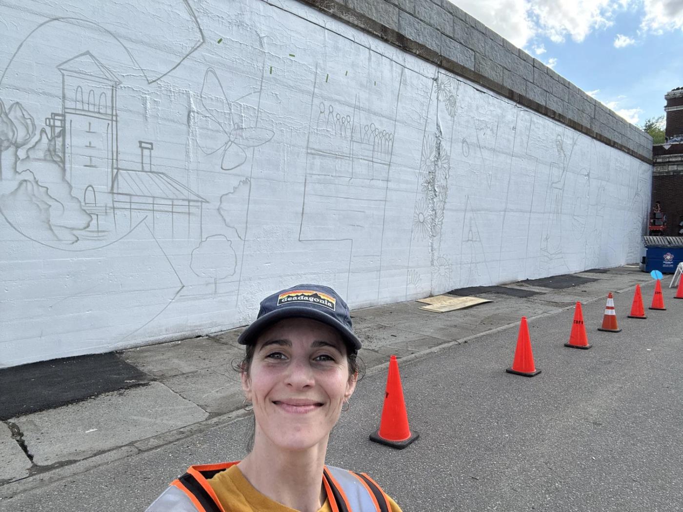 A woman, in a baseball cap and contruction vest, takes a selfie in front of a white wall with the outline of the mural drawn on.