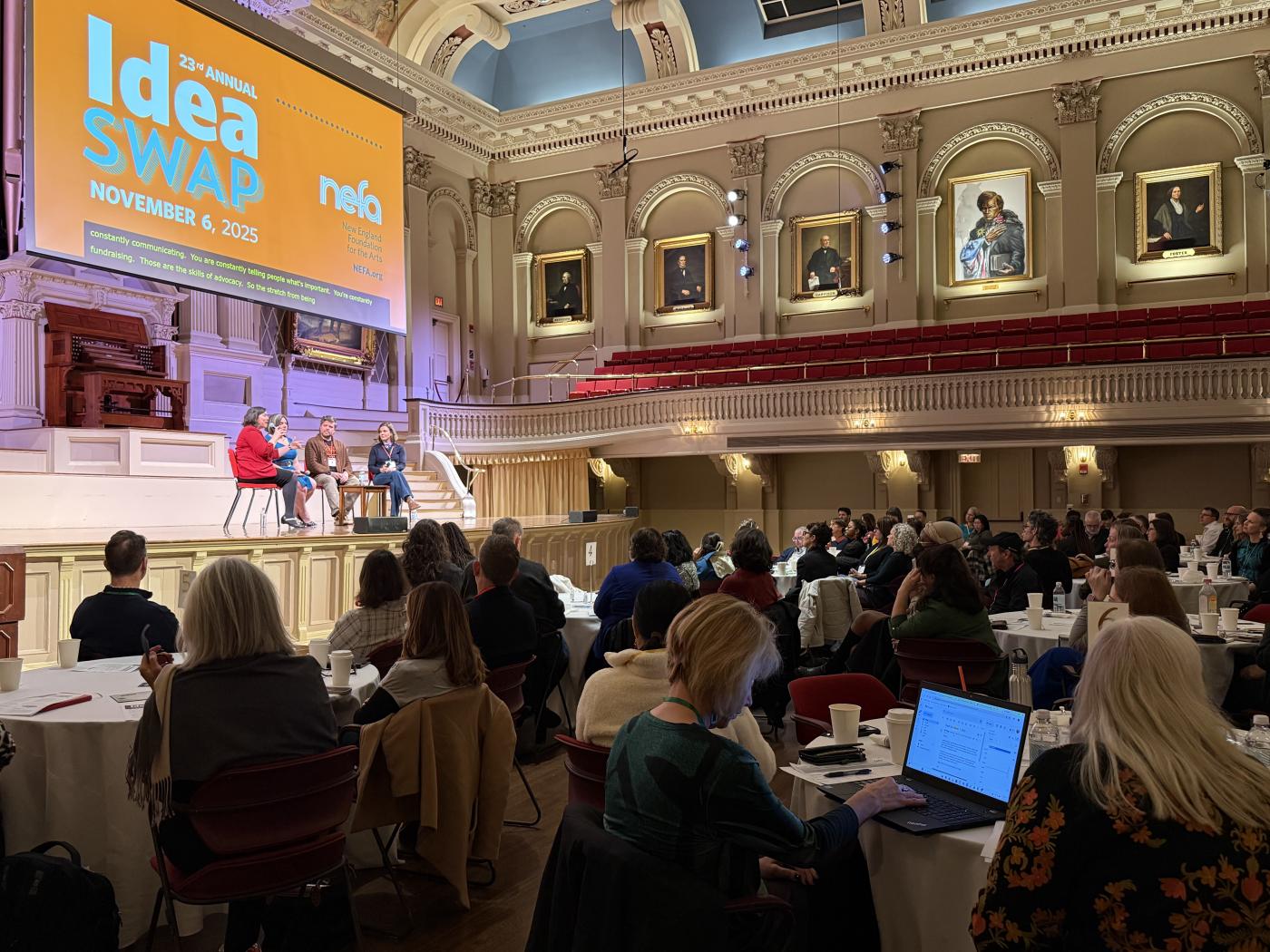 On a stage, with large event signage (Idea Swap 2025), four people sit in chairs before an audience of folks seated around tables.