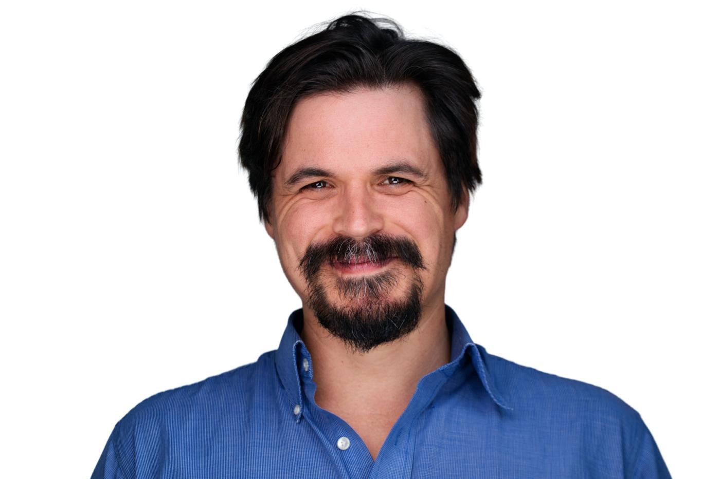 Chris Battaglia has dark brown hair and a goatee. He wears a denim shirt.