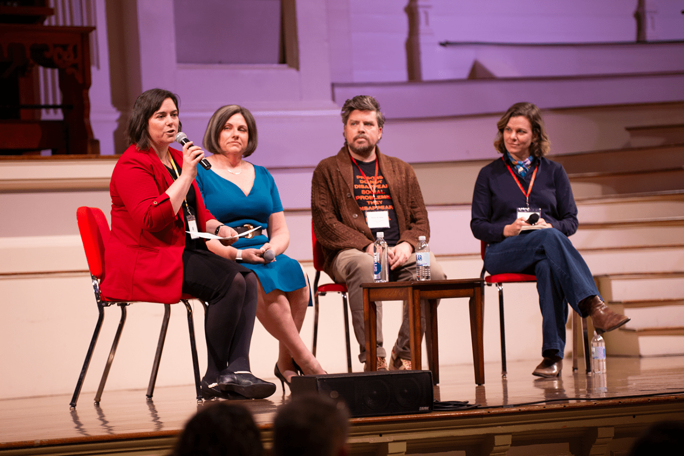 On a stage, a woman in a red blazer holds a microphone while three folks seated next to her watch.