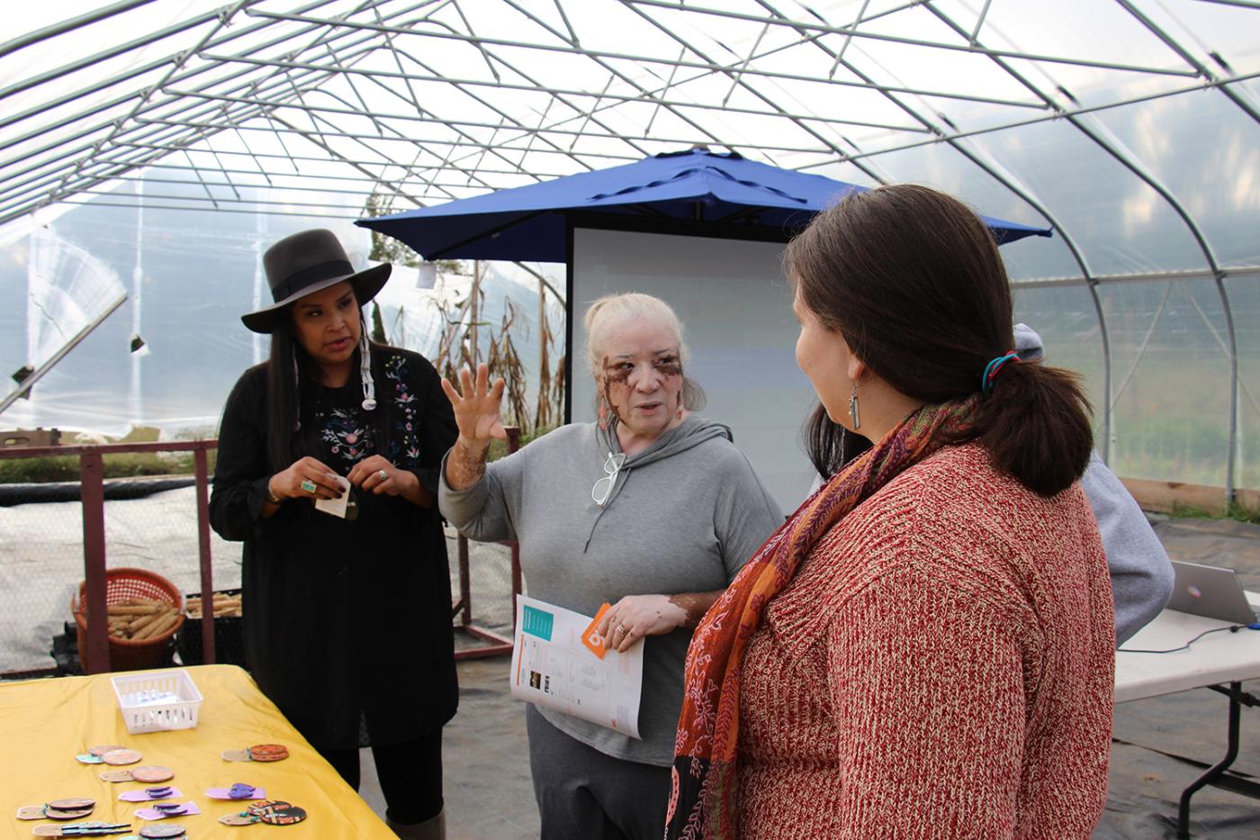Endawnis Spears (Diné/Ojibwe/Choctaw/Chicksaw), Gail Rokotuibau (Narragansett/Pequot), Samantha Fry (Narragansett), and Morganna Becker connect and chat together at the Markets & Marketing workshop for Native American artists inside the high tunnel. Endawnis’ jewelry is displayed on a table in the foreground.