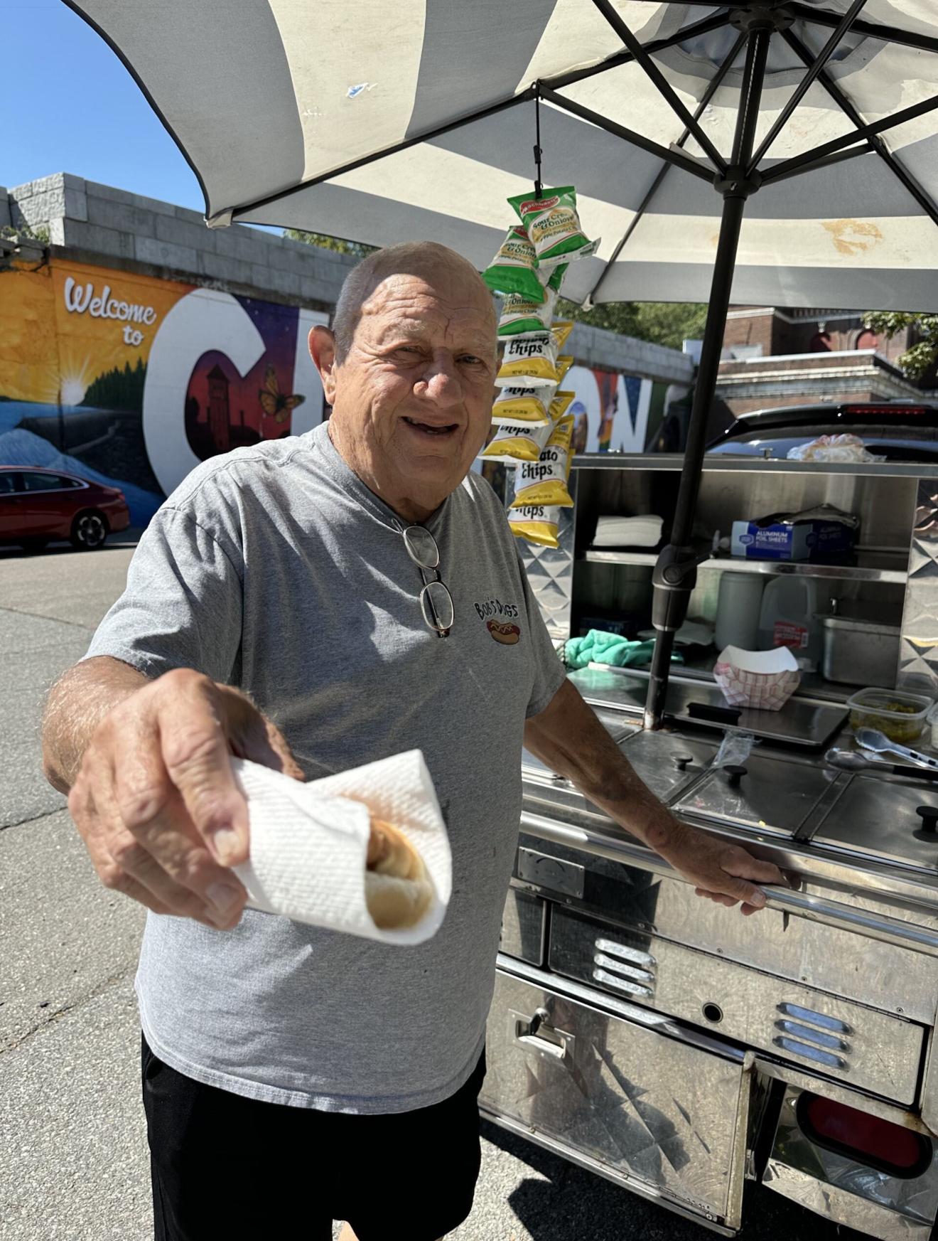 An elderly white man stands behind a hotdog stand. He hands the photographer a hotdog.
