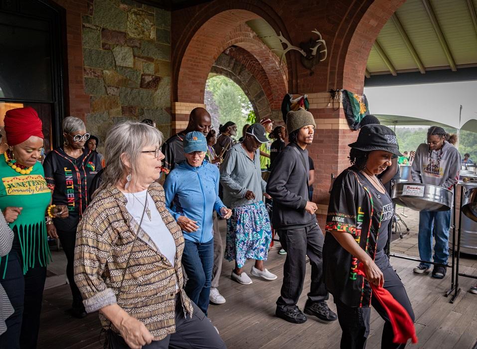 On a porch, a crowd of folks of various races follow the steps of a dancer who leads them.