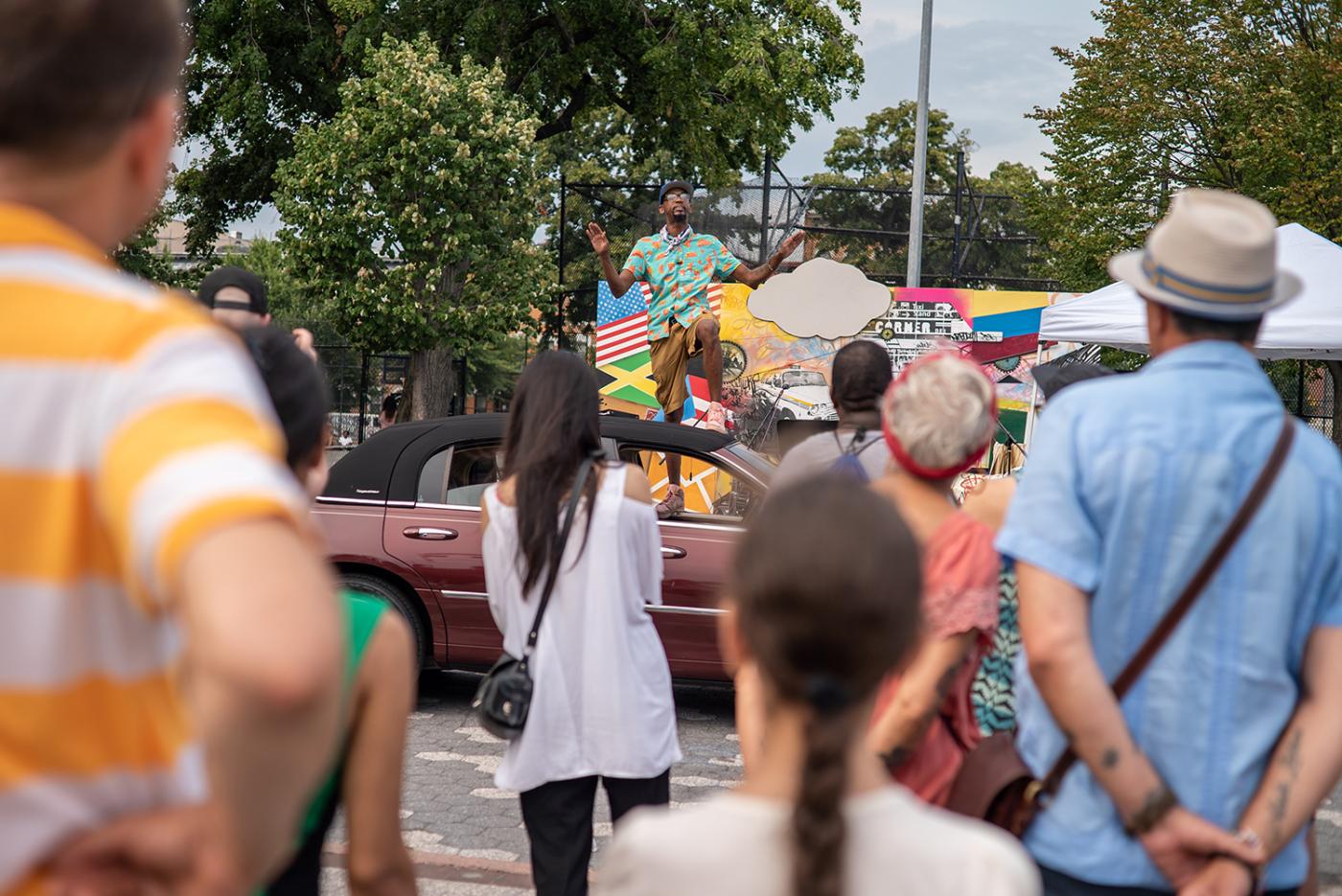 A Black man stands on a car and speaks to an audience in the foreground.