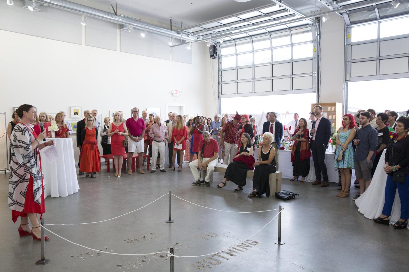 A crowd of people dressed in red and white watch a woman speak in a roped off area of a large, bright warehouse/garage