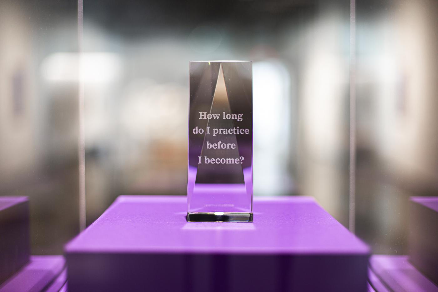 a trophy is displayed in a glass case with a purple bottom. the trophy reads "How long do I practice before I become?"