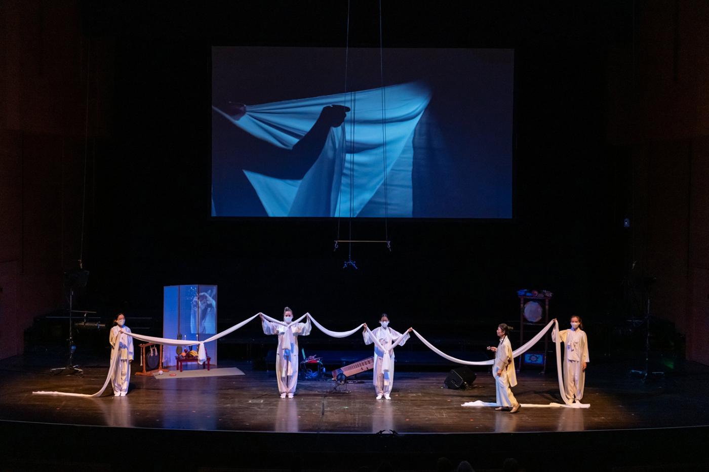On a stage, four folks, and Jin Hi, hold up a white cloth. behind them, a projection of a person with a white cloth.