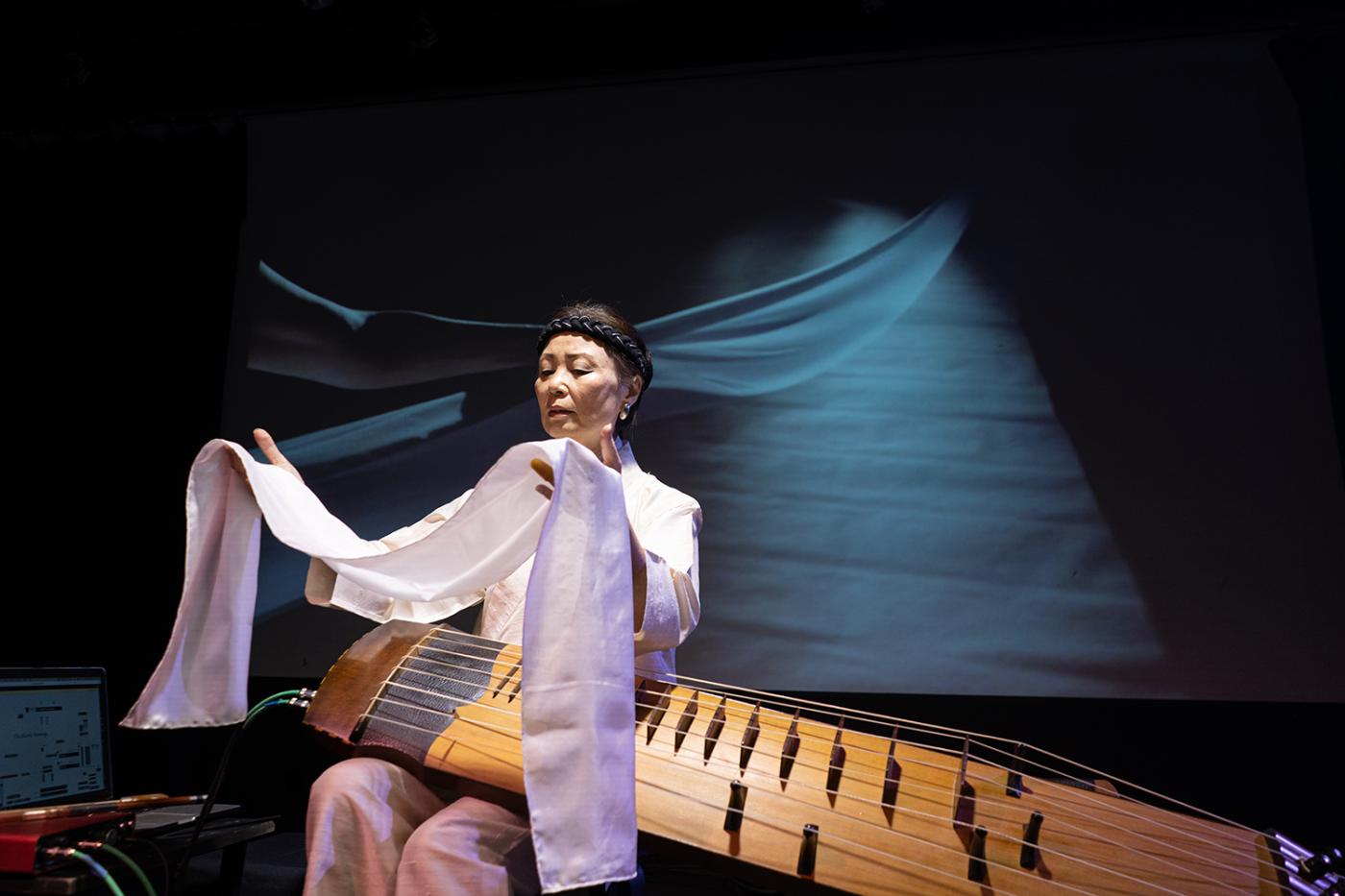 In stage lighting, Jin Hi holds up a white cloth over her komungo instrument.