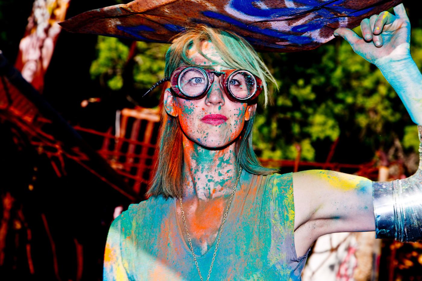 A woman wearing giant goggles is covered in colorful splattered paints
