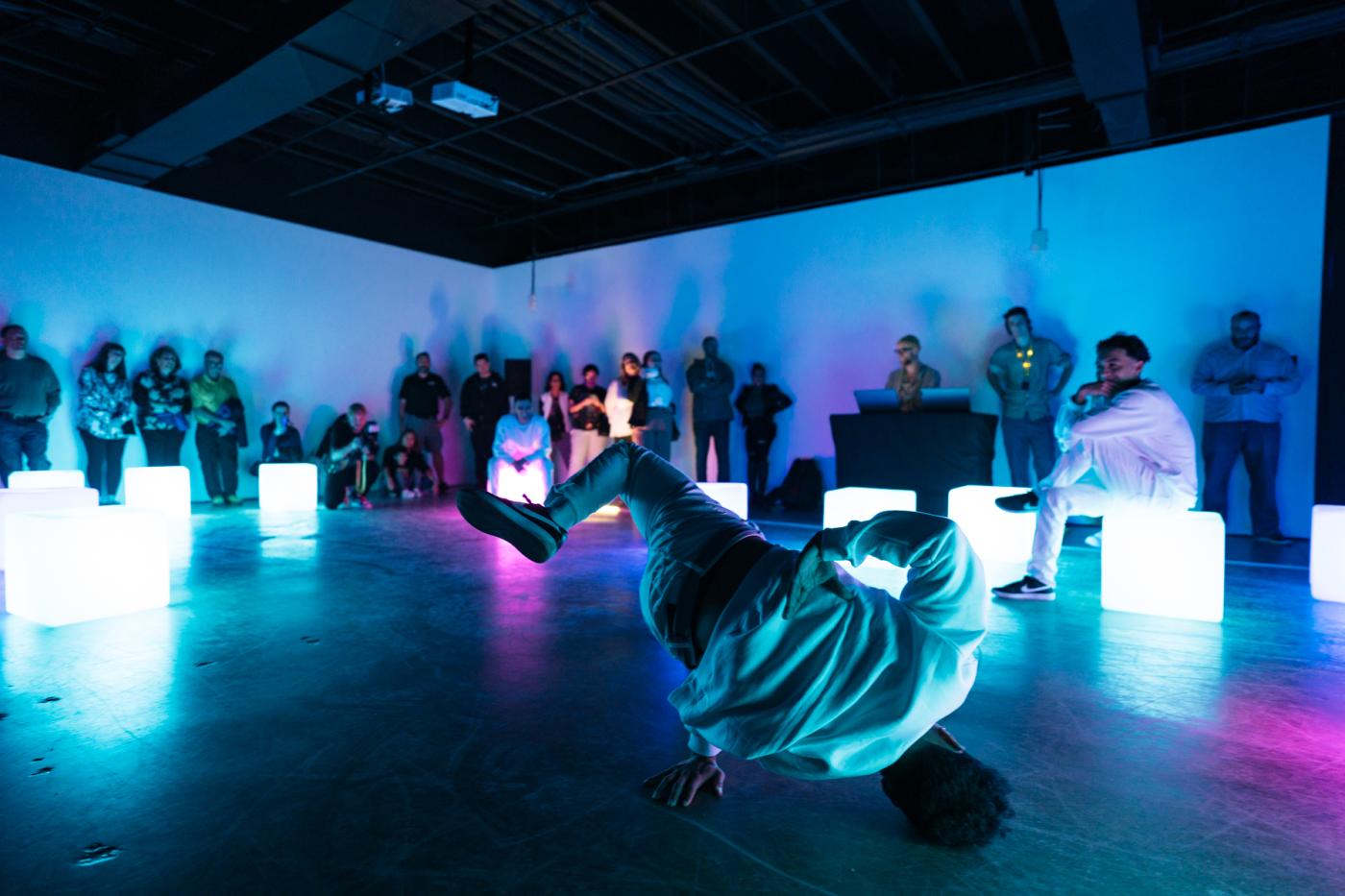 A person break dances in a room lined with square boxes that light up and an audience that is standing up against the walls beyond those boxes.