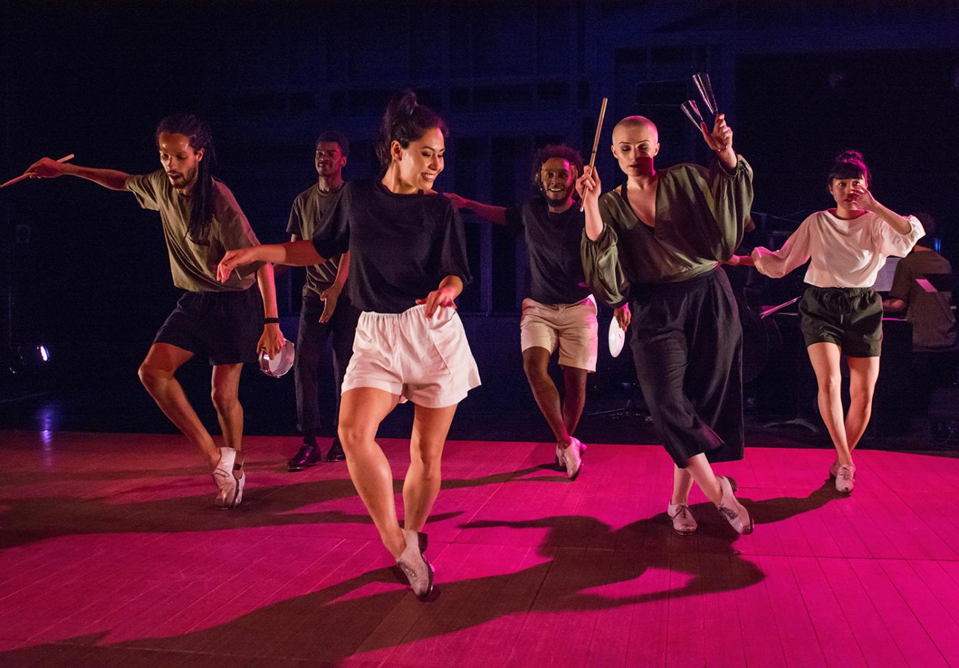 On a stage with a pink floor, six folks dance in casual outfits.