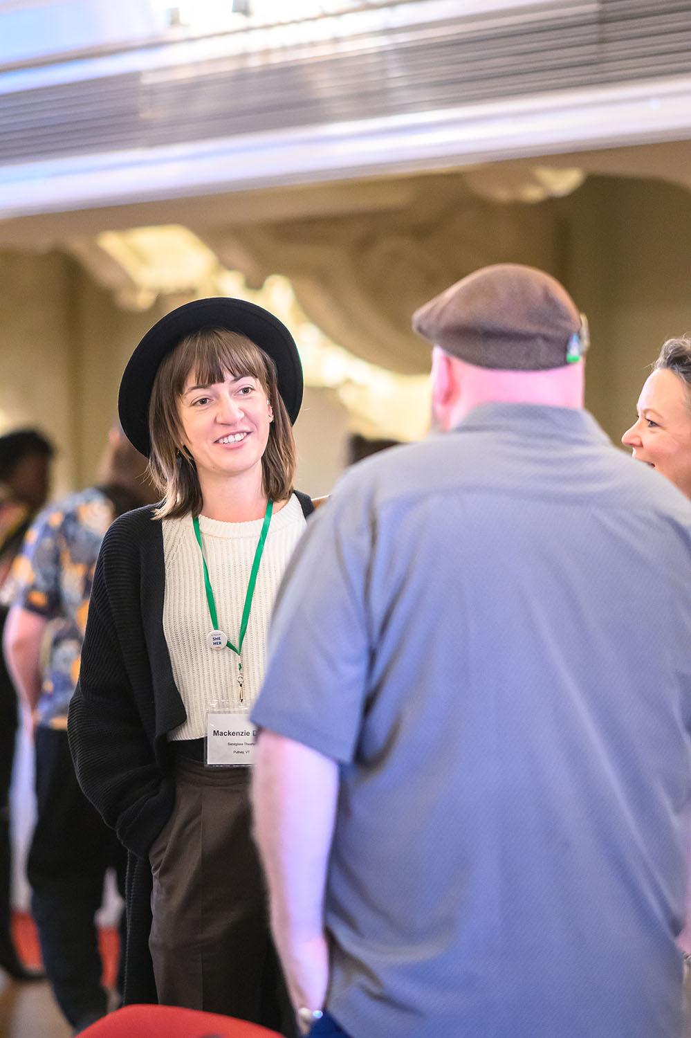 In a crowded space, a woman, in a black top hat, smiles at a man she's in conversation with. She wears a nametag: Mackenzie.
