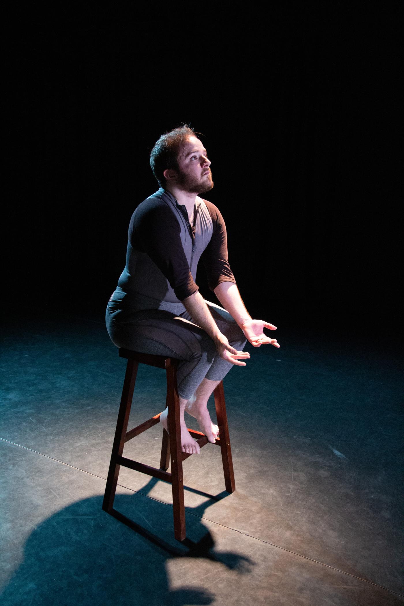 In a spotlight, Toby sits on a stool with their hands out.