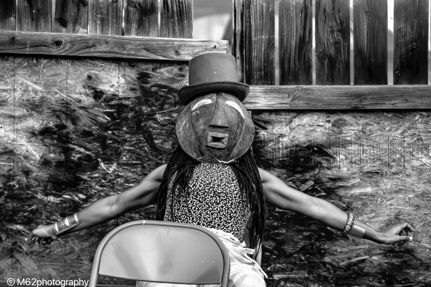In black and white, a Black woman wears a mask and a fedora.