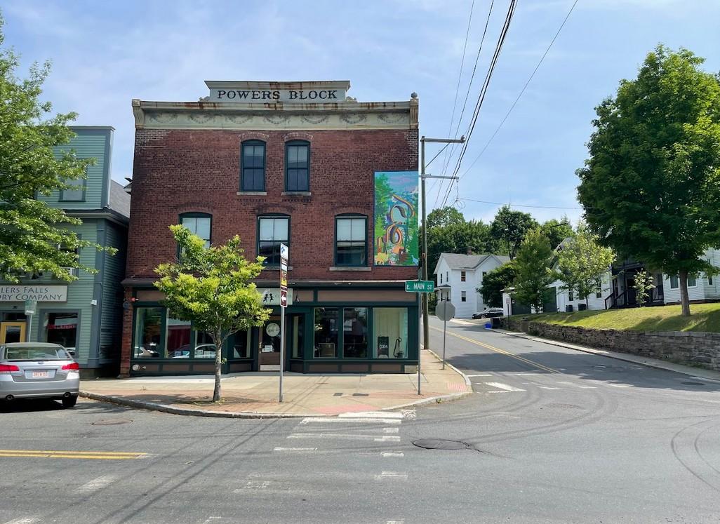 On a street corner, in a small town with no traffic or pedestrians, a mural on the side of a brick building. Below, the building has an awning. Street lines and trees frame the photo.