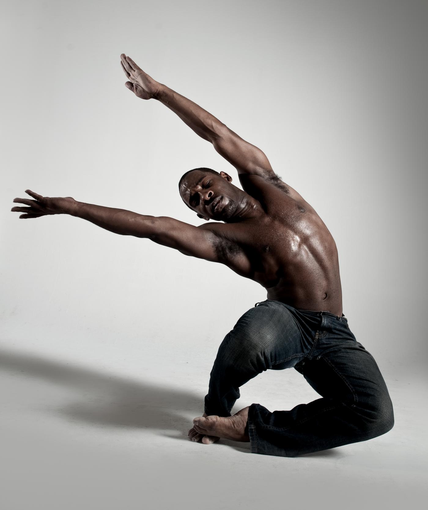 A male dancer holds his arms up and sideways, while squatting.