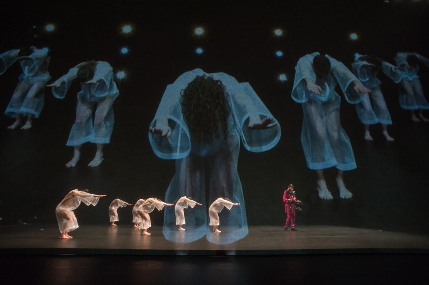 On a stage, six dancers in white and one in read perform beneath a projection of women floating in bell sleaved dresses.