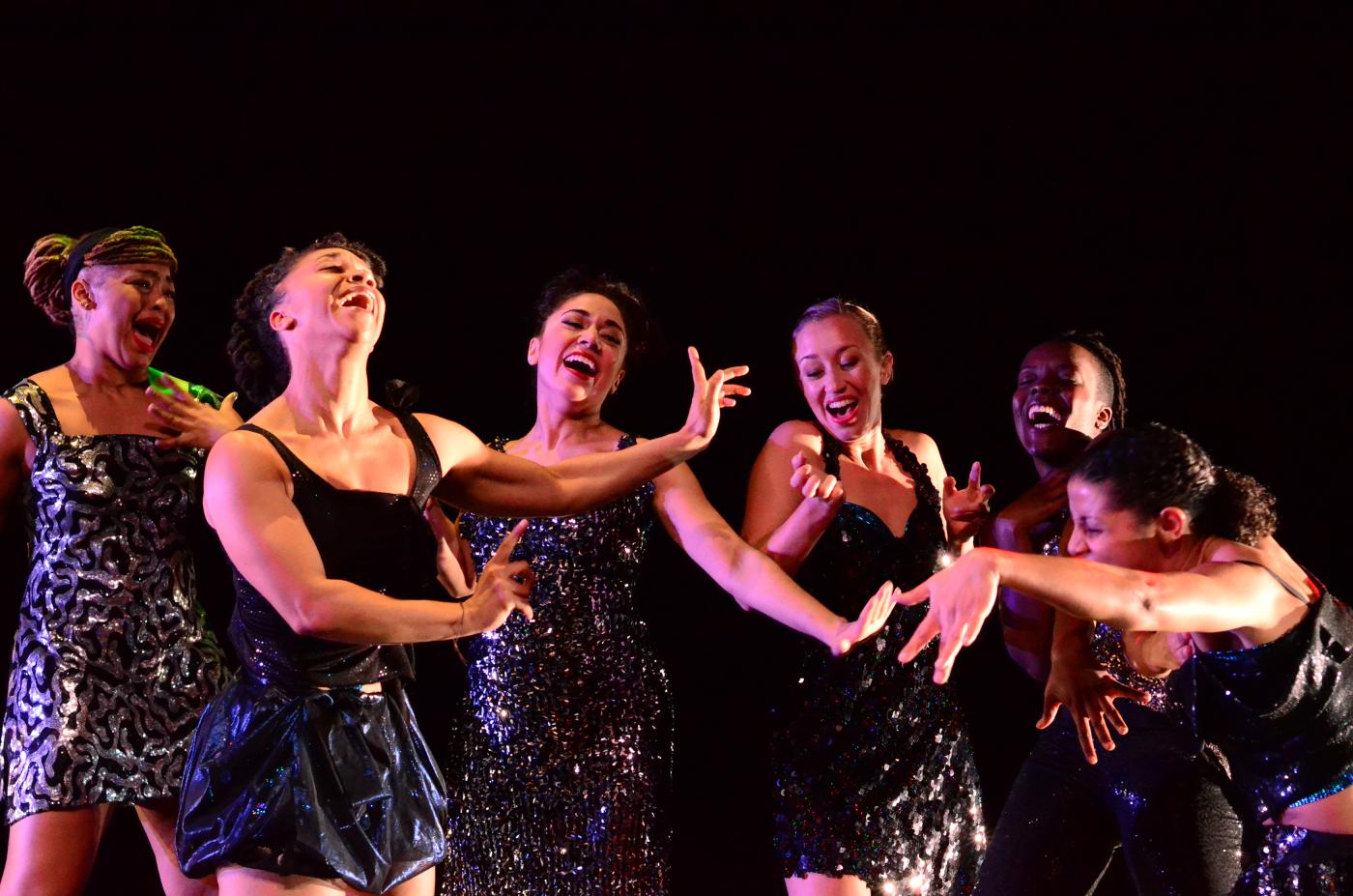 On a stage, a group of women laugh and move their arms, while wearing sequin dresses.