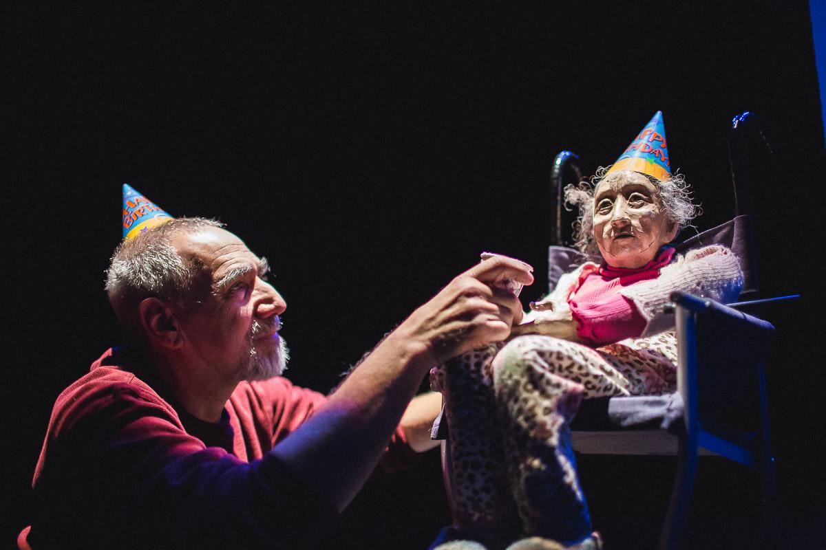 An older white man crouches next to a puppet of an elderly white woman in a wheel chair. They both wear conical birthday hats.