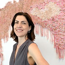 Nicole has brown hair and white or light skin. She poses in fro.nt of yarn art and wears a grey tank top