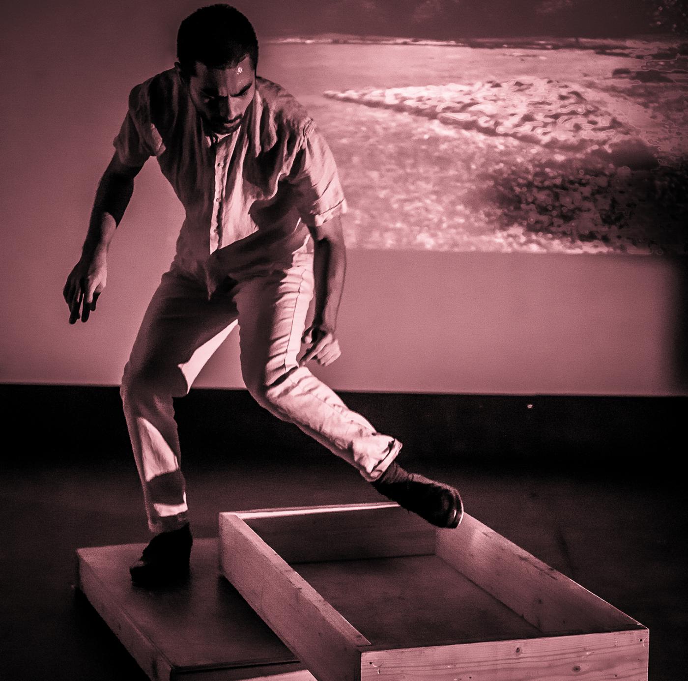 In front of a projection of the ocean, Orlando hops out of a small wooden frame on the floor.