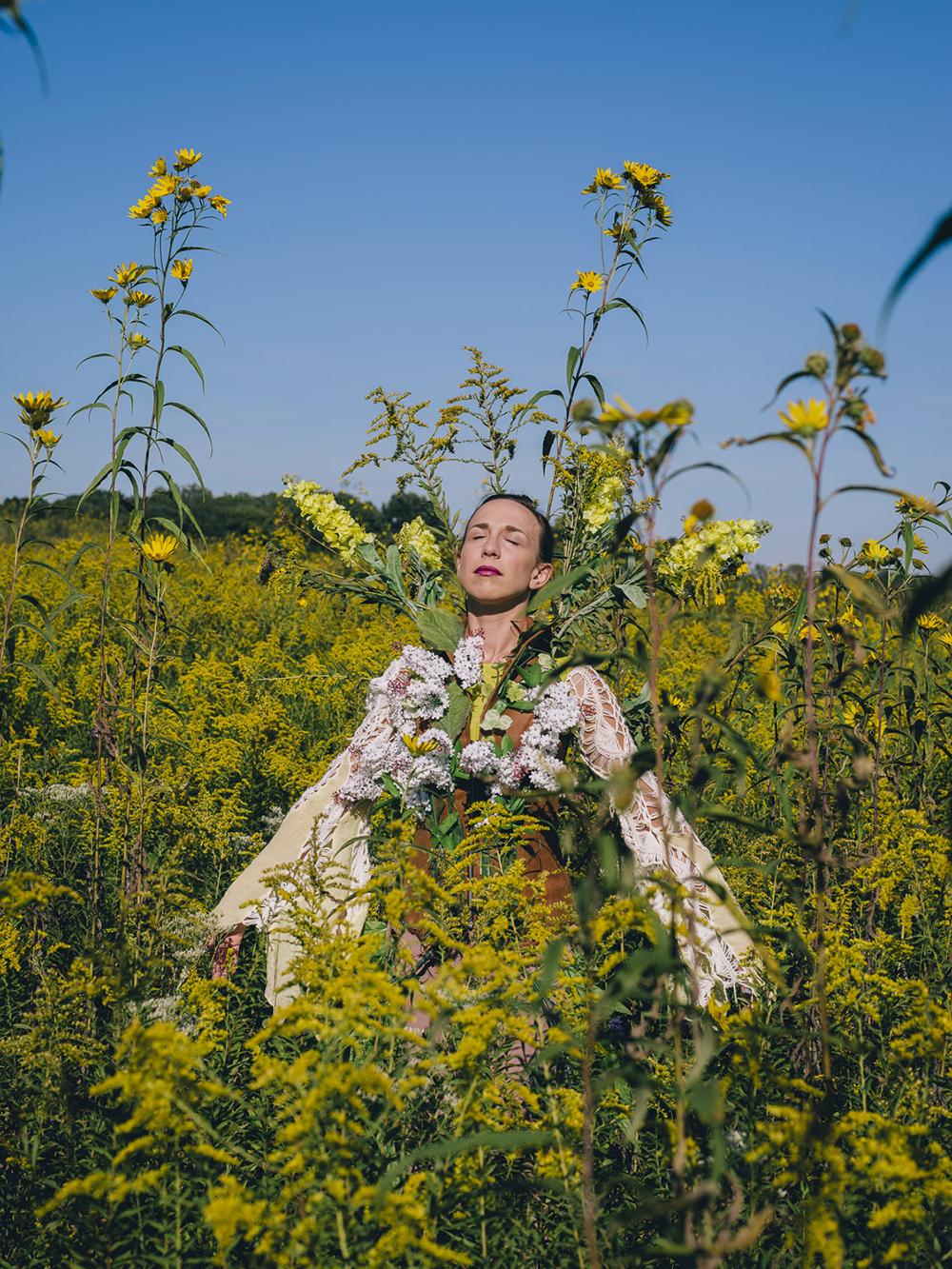 A white woman stands in field, intertwined with tall flowers.