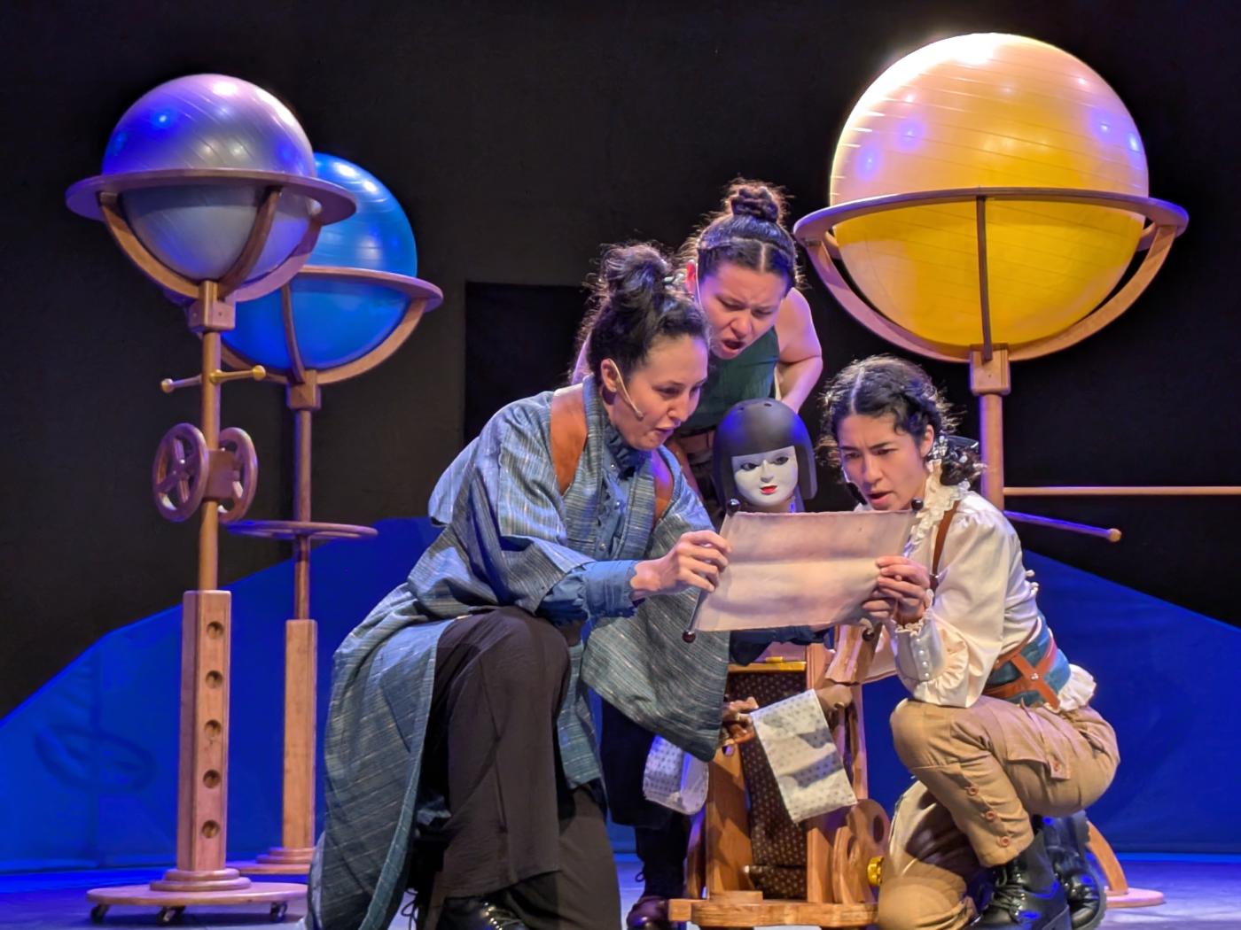 On a stage, with globes of different planets behind them, three girls and a puppet look at a piece of paper.