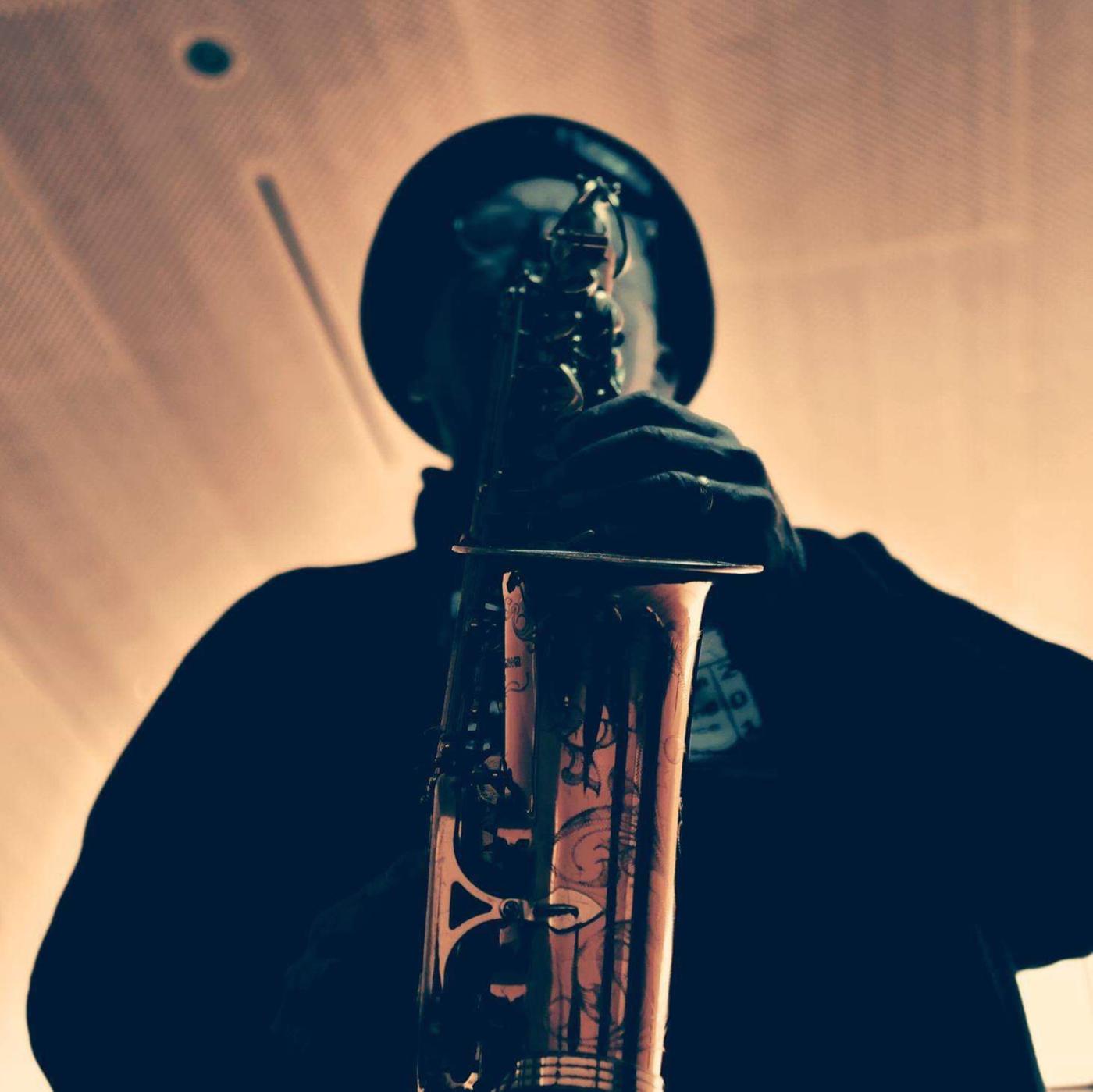 A black man plays the saxophone. His face is blocked by the instrument.