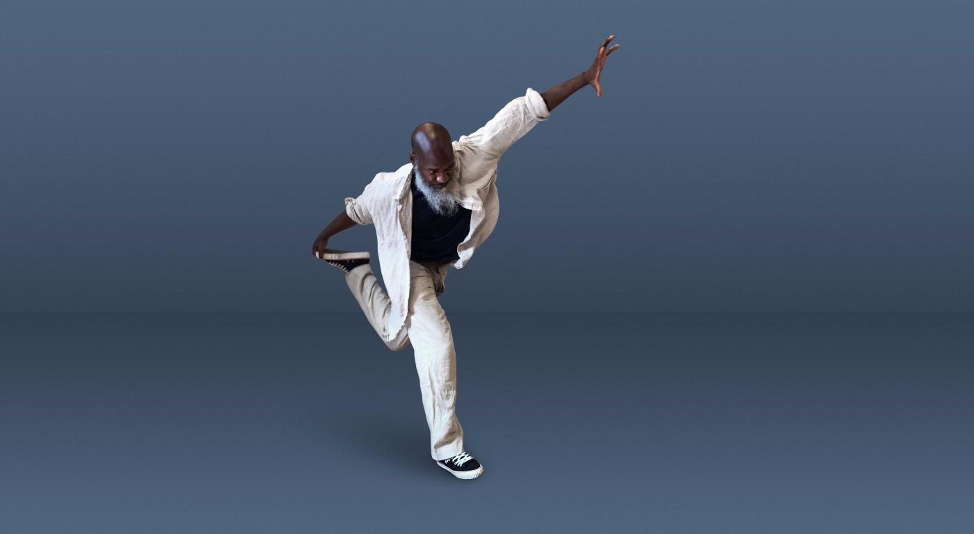 In photo studio and wearing a white cotton jacket and pants, Xavier holds his left arm up and his back leg up behind him.
