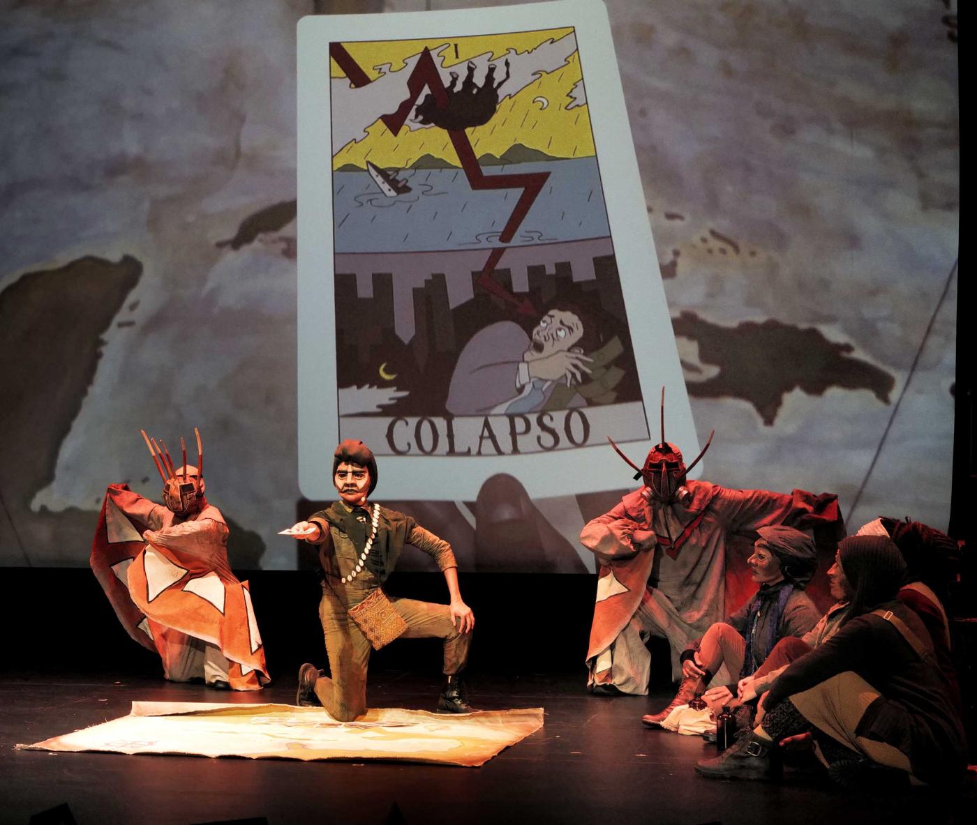 Actors wear masks and costumes. Projected behind them is a card that reads "colapso."