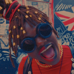 A Black woman with braids, and wearing goggles and red and blue lipstick on the left and right halves of her mouth, smirks.