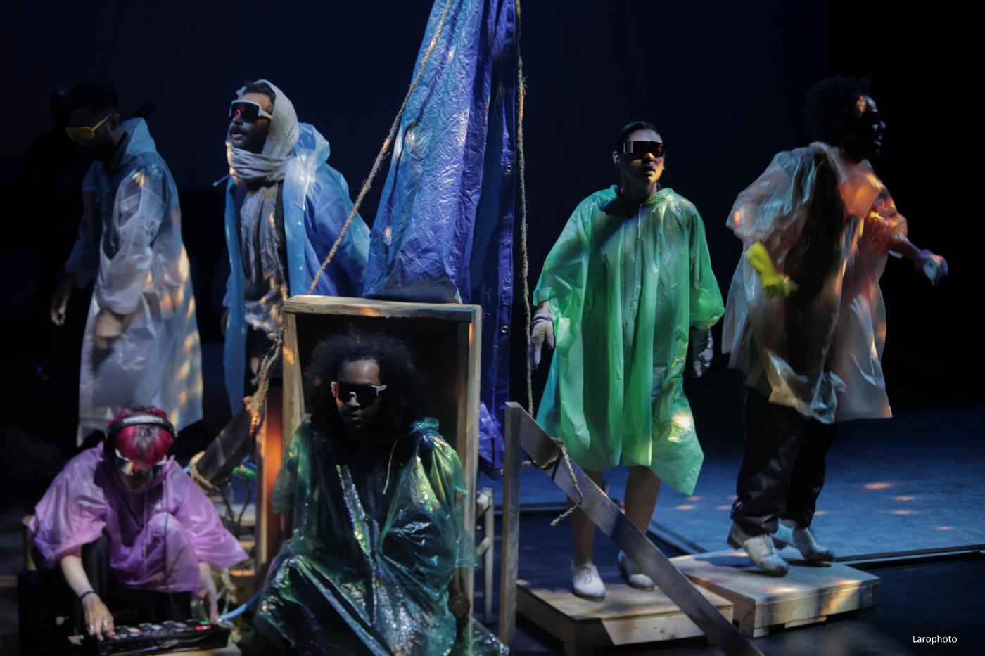 Six performers in colorful plastic rain ponchos and sunglasses stand on a stage.