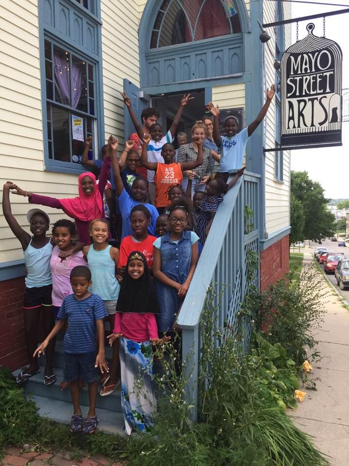 a smiling group of elementary school students pose on the deck of the Mayo Street Arts building