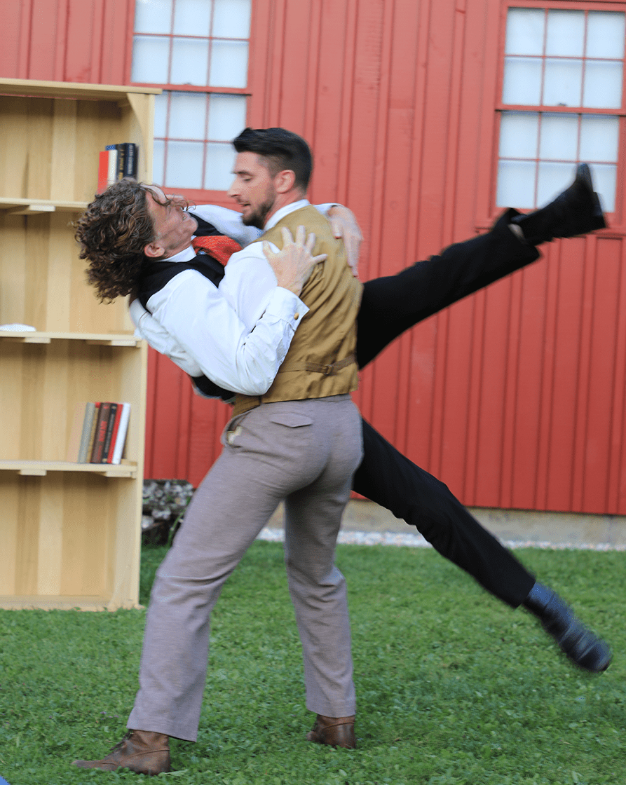 Outside, by a barn and a book shelf, two men in suits dance.