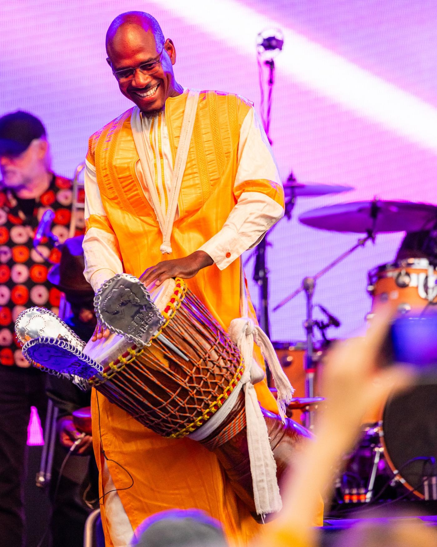 On a stage, a Black man plays the djembe and smiles for an audience that is blurry in the foreground.