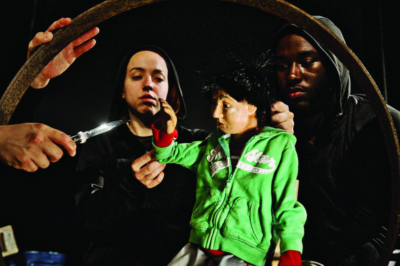 A puppet in a green hoodie, controlled by two more puppeteers, faces a small knife held by another person.