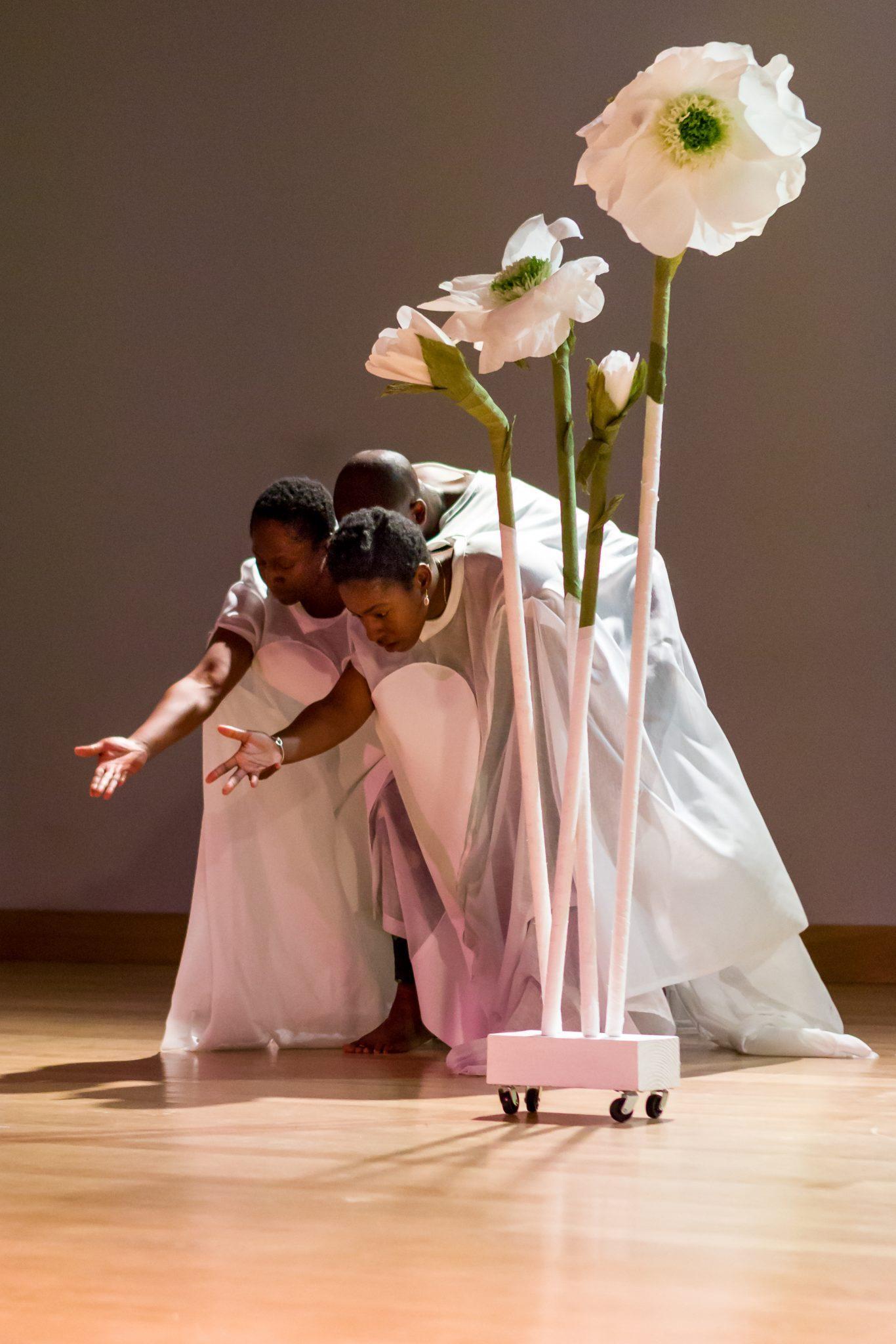 Three dancers perform next to very large, human-sized flowers.