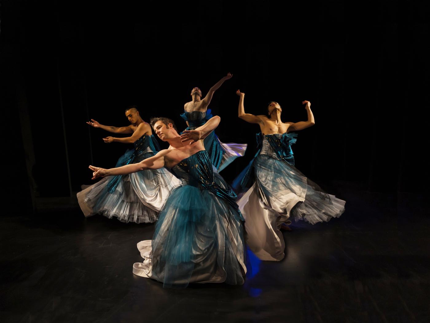 In a dim studio space, a group of men in blue-ish dresses dance in a circle.
