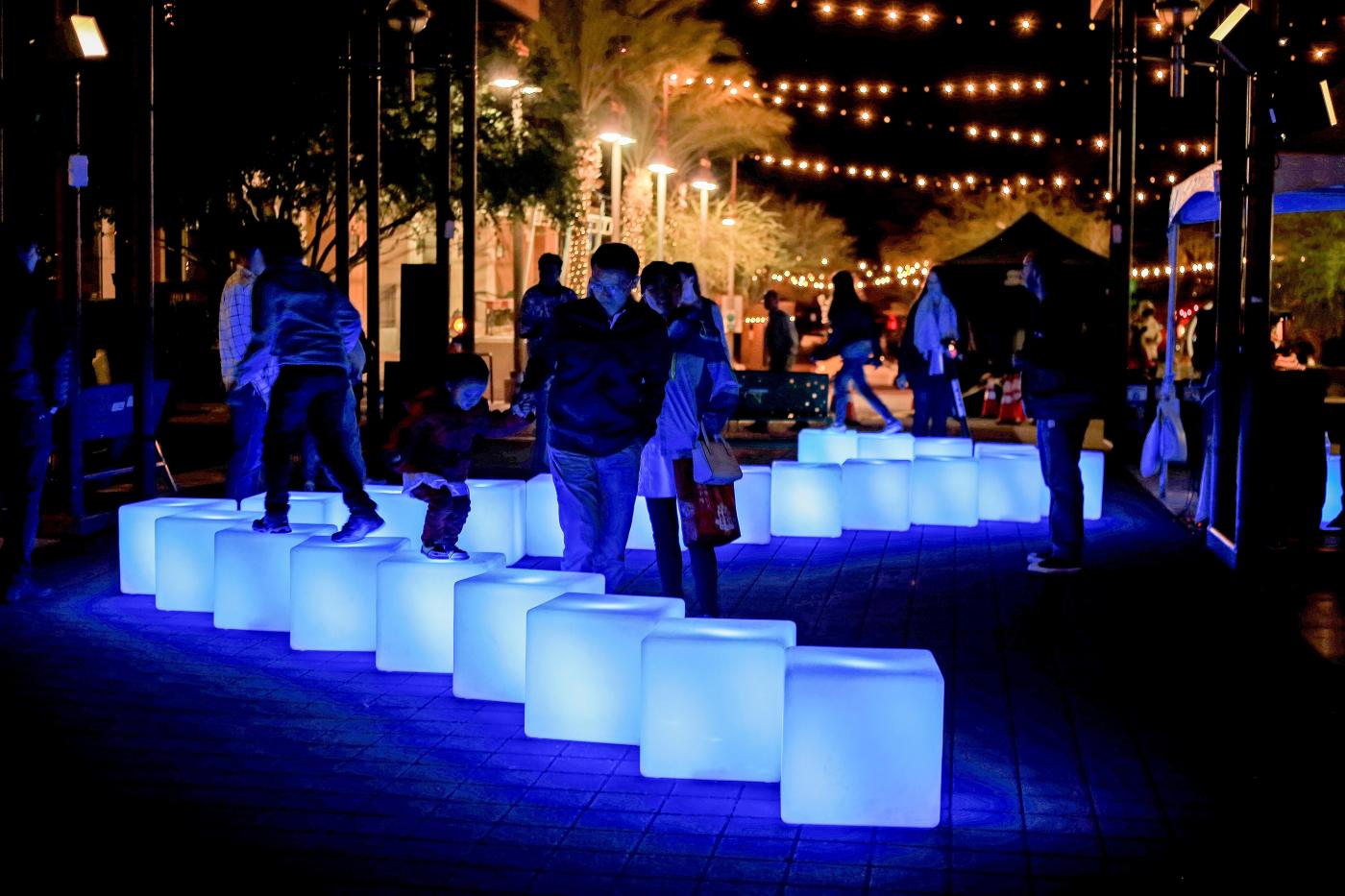 In a dark, city park, folks walk next to and on top of blocks that light up.