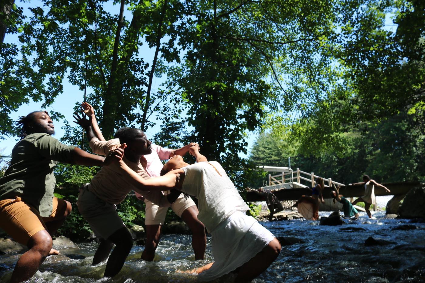 In a rocky river, eight dancers lean in different directions, supporting each other.