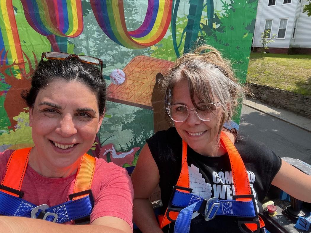 Two women sit in front of a similar rainbow motif to the mural with the rainbows. They have neon orange staps on their shoulders.