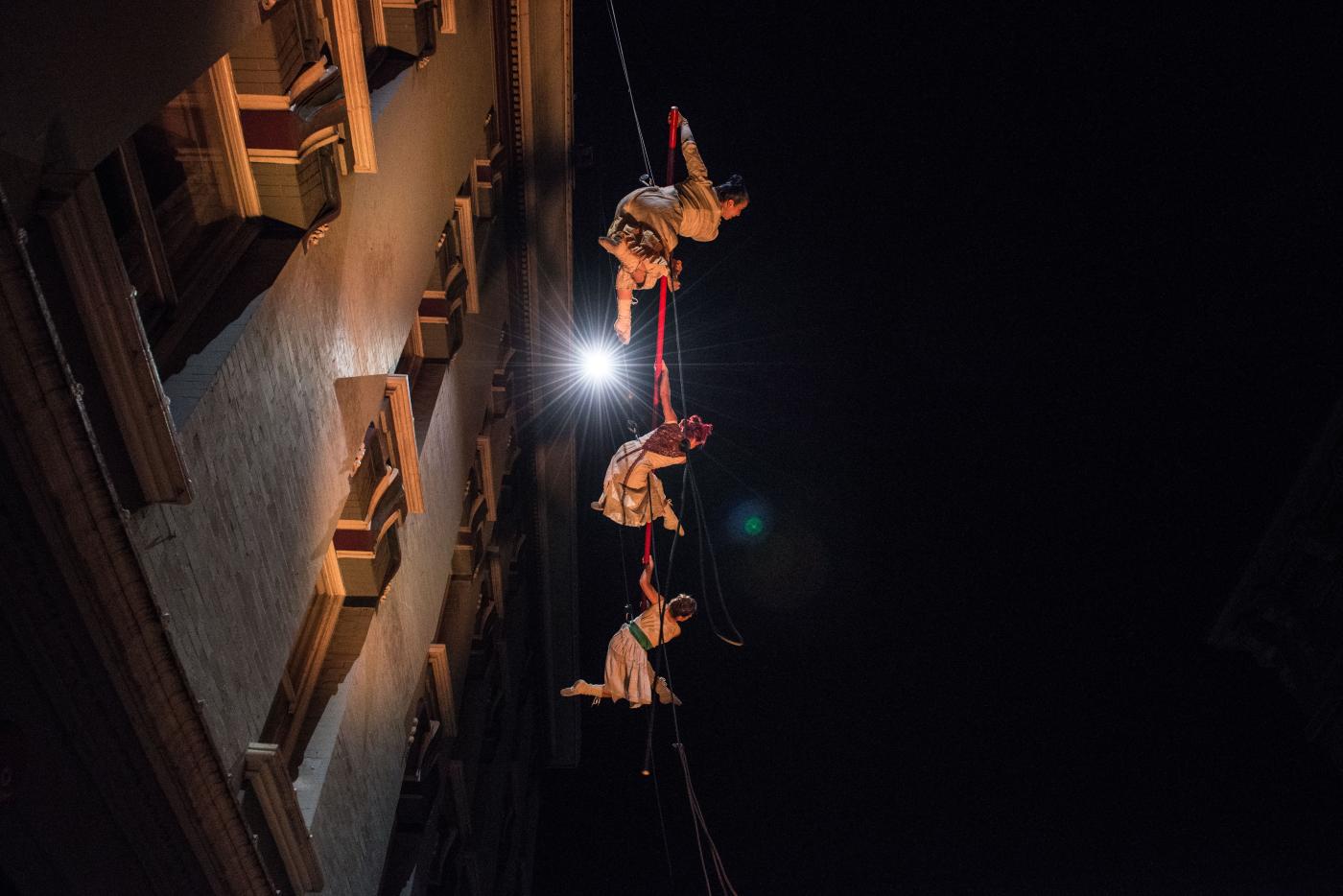 At night and in spotlights, three performers hang off the side of a building.