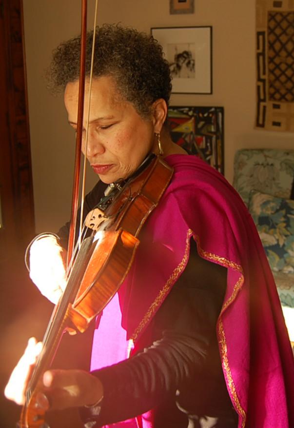 Terry is a woman of color. She wears a pink scarf on her shoulder below the violin she plays.