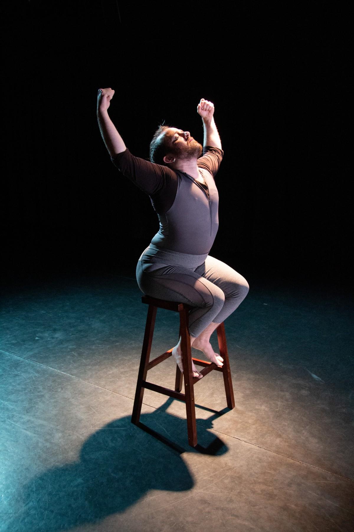 Toby in spotlight seated on a stool, back arched, arms raised but bent, fists clenched, head tilted back